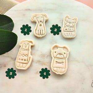 Puede incluir: Cuatro cortadores de galletas beige con forma de perro, cada uno con dise&ntilde;os &uacute;nicos, se muestran sobre una superficie con patr&oacute;n de m&aacute;rmol. Adhesivos florales verdes acompa&ntilde;an a los cortadores. La imagen tiene una est&eacute;tica suave y natural, ideal para hornear.