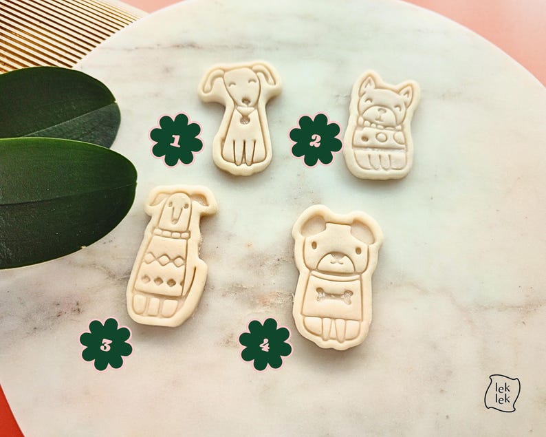 Puede incluir: Cuatro cortadores de galletas beige con forma de perro, cada uno con dise&ntilde;os &uacute;nicos, se muestran sobre una superficie con patr&oacute;n de m&aacute;rmol. Adhesivos florales verdes acompa&ntilde;an a los cortadores. La imagen tiene una est&eacute;tica suave y natural, ideal para hornear.
