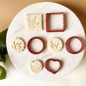 Puede incluir: Conjunto de siete cortadores de galletas rojos en varias formas, incluyendo un corazón, un círculo, un cuadrado y dos formas de manos. Los cortadores están dispuestos sobre una superficie blanca.