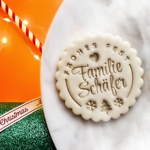 May include: A white cookie with a brown imprint that reads "Frohes Fest Familie Sch&auml;fer" with a heart and snowflakes.