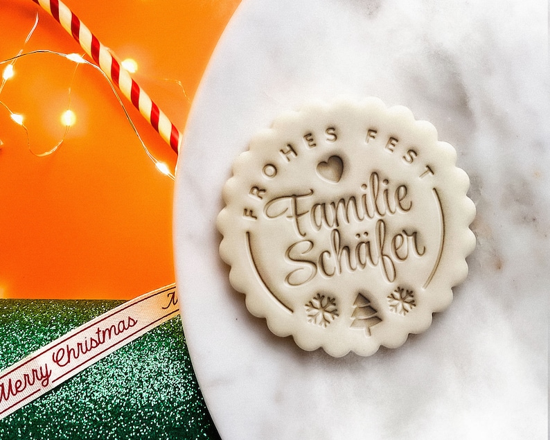 May include: A white cookie with a brown imprint that reads "Frohes Fest Familie Sch&auml;fer" with a heart and snowflakes.