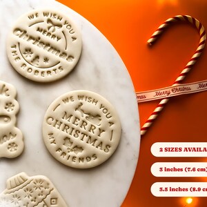 May include: Four white cookie dough rounds with different Christmas-themed designs. The designs include "We Wish You A Merry Christmas The Roberts", "We Wish You A Merry Christmas My Friends", "Think You Merry Christmas", and a Christmas ornament. The dough rounds are on a white marble surface. A red and white candy cane and a white ribbon with the text "Merry Christmas" are on a bright orange surface. The text "2 SIZES AVAILABLE - 3 inches (7.6 cm) 3.5 inches (8.9 cm)" is below the candy cane and ribbon.