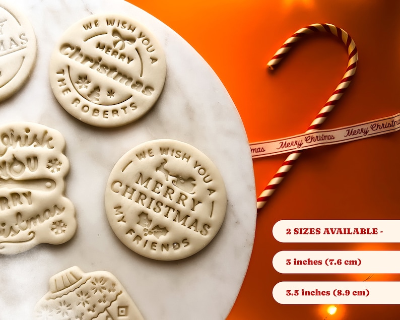 May include: Four white cookie dough rounds with different Christmas-themed designs. The designs include "We Wish You A Merry Christmas The Roberts", "We Wish You A Merry Christmas My Friends", "Think You Merry Christmas", and a Christmas ornament. The dough rounds are on a white marble surface. A red and white candy cane and a white ribbon with the text "Merry Christmas" are on a bright orange surface. The text "2 SIZES AVAILABLE - 3 inches (7.6 cm) 3.5 inches (8.9 cm)" is below the candy cane and ribbon.