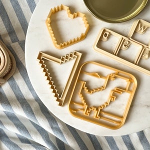 Könnte beinhalten: Vier Ausstecher auf einer weißen Marmorfläche. Die Ausstecher sind in Form eines Herzens, eines pixelartigen Pfeils, eines pixelartigen Vogels und einer Tastatur mit den Buchstaben W, A, S und D.