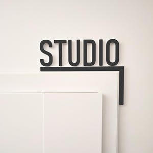 Puede incluir: Letrero negro con la palabra "STUDIO" en negrita, montado en una pared blanca y sobre un marco de puerta blanco. El letrero y la tipografía crean un aspecto moderno y minimalista, adecuado para un espacio creativo.