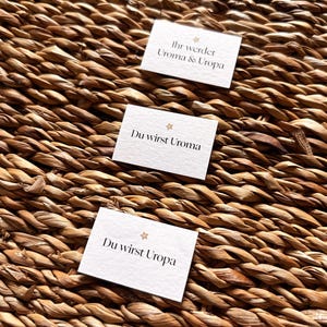 May include: Three small white cards with German text are arranged on a woven, brown surface. The cards read "Ihr werdet Uroma & Uropa", "Du wirst Uroma", and "Du wirst Uropa". Each card has a small star design.