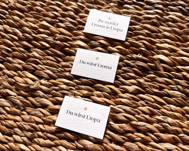 May include: Three small white cards with German text are arranged on a woven, brown surface. The cards read "Ihr werdet Uroma & Uropa", "Du wirst Uroma", and "Du wirst Uropa". Each card has a small star design.
