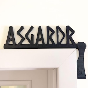Op de afbeelding: Zwart houten bord met het woord "Asgard" in een gestileerd lettertype, met een bijl-vormig handvat aan de rechterkant. Het bord is gemonteerd boven een witte deur.