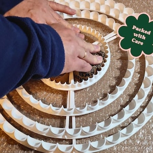 May include: A white, multi-tiered, scalloped-edge craft ring is being handled. The ring is made of plastic and has a circular design. The image includes a green flower-shaped sticker that says "Packed with Care".
