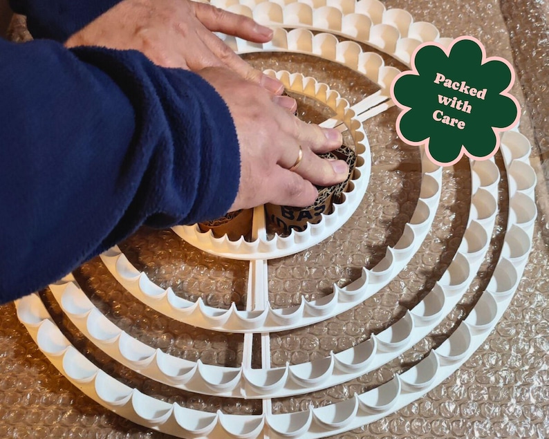 May include: A white, multi-tiered, scalloped-edge craft ring is being handled. The ring is made of plastic and has a circular design. The image includes a green flower-shaped sticker that says "Packed with Care".