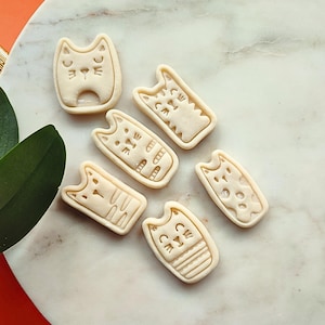 May include: A collection of seven beige cat-shaped cookie cutters. Each cutter features a different cat face design, with details like eyes, whiskers, and stripes. The cutters are arranged on a marble surface.