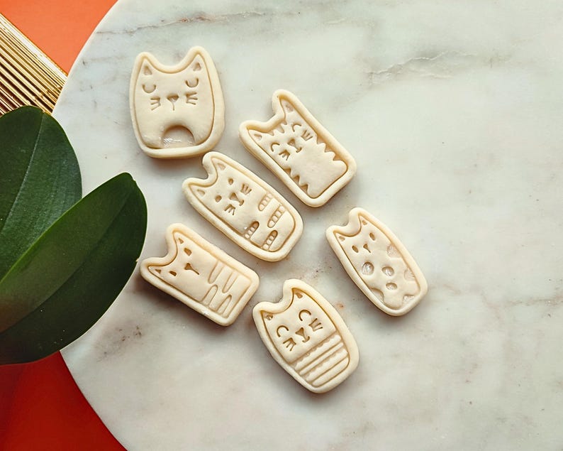 May include: A collection of seven beige cat-shaped cookie cutters. Each cutter features a different cat face design, with details like eyes, whiskers, and stripes. The cutters are arranged on a marble surface.