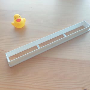 Op de afbeelding: Een witte rechthoekige koekjessnijder met twee interne steunen ligt op een licht houten oppervlak. Naast de uitsteker staat een kleine gele badeend met een oranje snavel. De eend kijkt naar links.