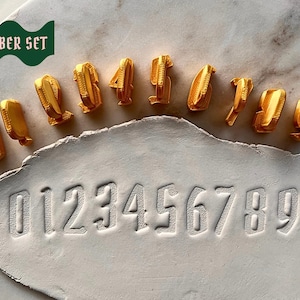 May include: A set of ten gold-colored number stamps from zero to nine, arranged in a curved line above a white clay slab. The clay slab features the embossed impressions of the numbers zero through nine in a clear, sans-serif font style.