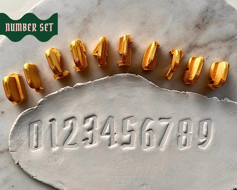 May include: A set of ten gold-colored number stamps from zero to nine, arranged in a curved line above a white clay slab. The clay slab features the embossed impressions of the numbers zero through nine in a clear, sans-serif font style.