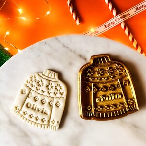 May include: Two Christmas cookie cutters in the shape of sweaters. The cutter on the left is white and says "Olivia" on the front. The cutter on the right is gold and says "Willow" on the front.