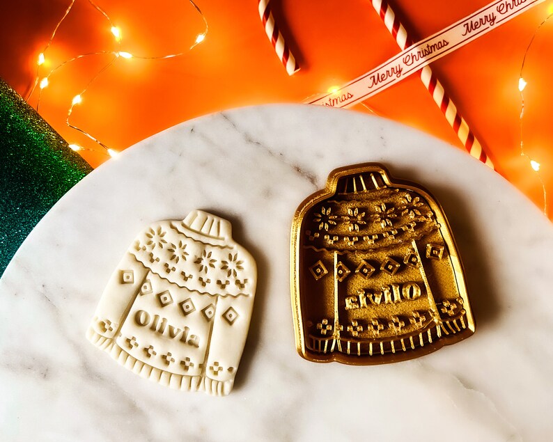 May include: Two Christmas cookie cutters in the shape of sweaters. The cutter on the left is white and says "Olivia" on the front. The cutter on the right is gold and says "Willow" on the front.