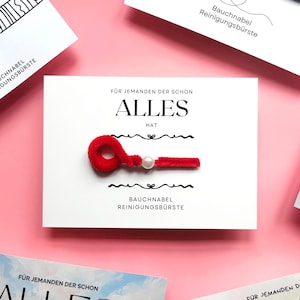 May include: A white card with German text and a red, fuzzy cleaning tool with a pearl. The card reads "FÜR JEMANDEN DER SCHON ALLES HAT" and "BAUCHNABEL REINIGUNGSBÜRSTE". Other cards are visible on a pink background.