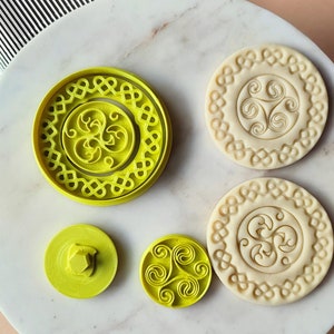May include: Four cookie cutters, two are white with a Celtic knot design and two are yellow with a Celtic knot design. The yellow cutters are different sizes.