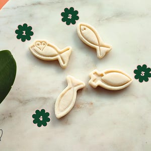 May include: Four beige fish-shaped cookie cutters on a marble surface. One cutter has a heart design. The cutters are arranged with green flower stickers. The image has a natural, bright aesthetic.