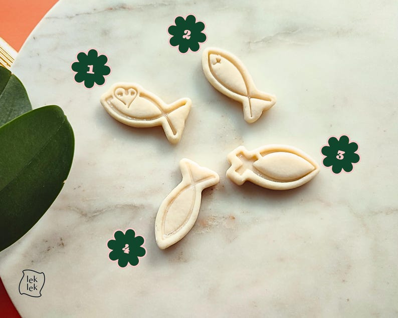 May include: Four beige fish-shaped cookie cutters on a marble surface. One cutter has a heart design. The cutters are arranged with green flower stickers. The image has a natural, bright aesthetic.