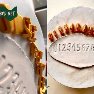 May include: A set of ten gold-colored 3D-printed number stamps from 0 to 9, arranged in a semi-circle on a white marble surface. Below the stamps, a white clay slab features the embossed sequence 0123456789. The text "NUMBER SET" is visible.