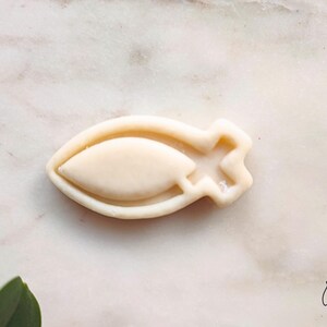 May include: A light beige fish-shaped cookie cutter. The cutter has a rounded body with a pointed end and a star-shaped tail. The cookie cutter is made of a light-colored material and is set against a white marble surface.