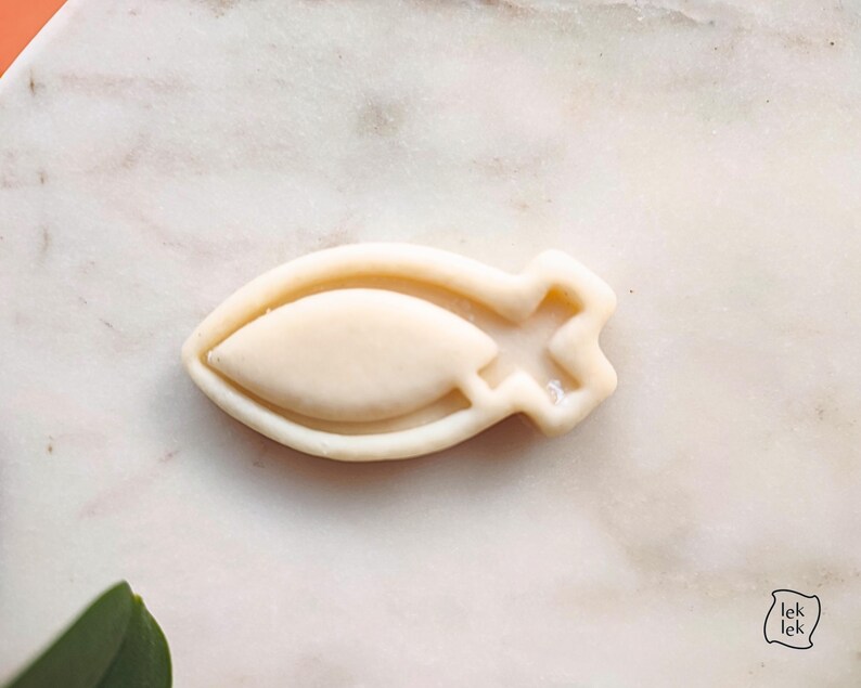 May include: A light beige fish-shaped cookie cutter. The cutter has a rounded body with a pointed end and a star-shaped tail. The cookie cutter is made of a light-colored material and is set against a white marble surface.
