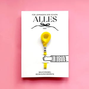 May include: A white card with the German text "F&Uuml;R JEMANDEN DER SCHON ALLES HAT" and a yellow fluffy object with the number "06" on it. The card also features a cartoon hand and the text "BAUCHNABEL REINIGUNGSB&Uuml;RSTE".