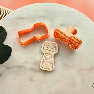 Puede incluir: Tres cortadores de galletas de pl&aacute;stico naranja: un zapato, un perro y una forma. El cortador de perro es un contorno detallado con manchas. El cortador de zapatos tiene un tal&oacute;n y una suela. El cortador de formas es un contorno simple.