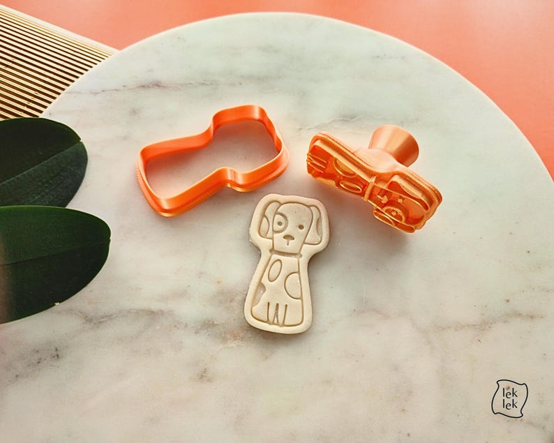 Puede incluir: Tres cortadores de galletas de pl&aacute;stico naranja: un zapato, un perro y una forma. El cortador de perro es un contorno detallado con manchas. El cortador de zapatos tiene un tal&oacute;n y una suela. El cortador de formas es un contorno simple.