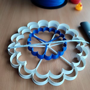 Op de afbeelding: Een witte, bloemvormige plastic mal met een blauwe, bloemvormige plastic mal erin. De mallen worden gemeten met een meetlint dat 17,8 cm aangeeft.