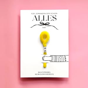 Op de afbeelding: Een witte rechthoekige kaart met de Duitse tekst F&Uuml;R JEMANDEN DER SCHON ALLES HAT en BAUCHNABEL REINIGUNGSB&Uuml;RSTE. Een gele pluizige pijpenrager in de vorm van een lus is bevestigd, voorzien van twee witte vierkante kralen met het getal 50.