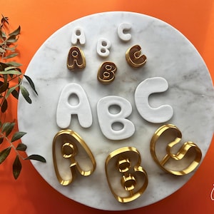 Puede incluir: Un juego de cortadores de galletas de metal dorado con las formas de las letras A, B y C, acompañados de recortes de arcilla o masa blanca en dos tamaños diferentes, dispuestos sobre una superficie redonda de mármol blanco con vetas grises.