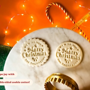 May include: Two white cookie dough rounds with the text "We wish you a Merry Christmas The Thompsons" embossed on them. A gold cookie cutter is in the foreground.