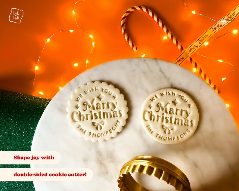 May include: Two white cookie dough rounds with the text "We wish you a Merry Christmas The Thompsons" embossed on them. A gold cookie cutter is in the foreground.