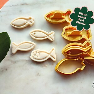 May include: A collection of fish-shaped cookie cutters and cookies. The cutters are yellow and the cookies are beige. The image includes the text "Plant-based PLA Plastic". The cookies have a cross or heart design.