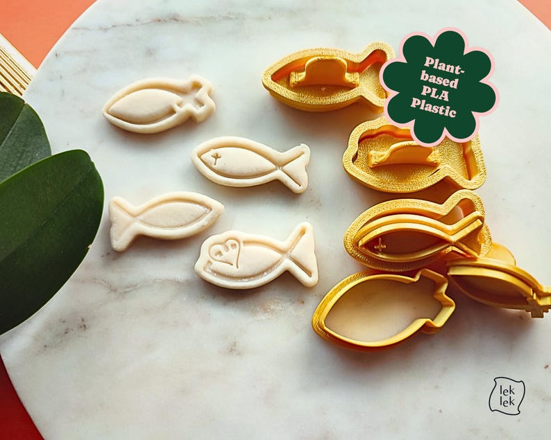 May include: A collection of fish-shaped cookie cutters and cookies. The cutters are yellow and the cookies are beige. The image includes the text "Plant-based PLA Plastic". The cookies have a cross or heart design.