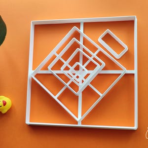 May include: White geometric cookie cutters in square and diamond shapes are arranged on an orange background. A small yellow rubber duck is in the lower left corner. The cutters are for baking and crafting.