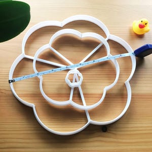 May include: White plastic flower-shaped cookie cutter with multiple layers. The cutter is 17.8 cm in diameter.