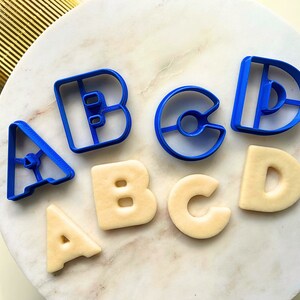 Pode incluir: Um conjunto de cortadores de biscoitos de plástico azul em forma de letras A, B, C e D, juntamente com quatro biscoitos cortados nas mesmas formas.