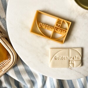 Pode incluir: Dois cortadores de biscoitos dourados, um com o texto "Golden Ratio" e o outro com um design geométrico. Os cortadores estão sobre uma superfície de mármore branco.