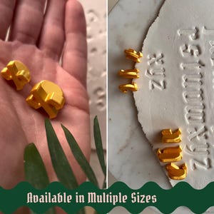 May include: A set of small, three-dimensional golden letter stamps used for embossing. The letters are shown in various sizes, with some held in a palm and others placed next to a white clay surface featuring impressed alphabet characters.