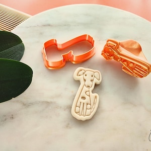 Puede incluir: Un cortador de galletas en forma de perro de color naranja y un sello a juego, junto a una galleta con forma de perro. La galleta es de color beige y tiene un dise&ntilde;o detallado de perro. El conjunto est&aacute; sobre una superficie de m&aacute;rmol.
