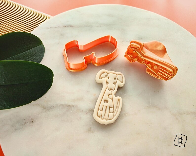 Puede incluir: Un cortador de galletas en forma de perro de color naranja y un sello a juego, junto a una galleta con forma de perro. La galleta es de color beige y tiene un dise&ntilde;o detallado de perro. El conjunto est&aacute; sobre una superficie de m&aacute;rmol.