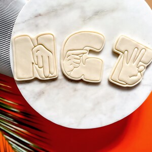May include: Three white cookie cutters shaped like hands forming the letters "NG7" in American Sign Language.