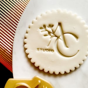 Könnte beinhalten: Weißer Tonstempel mit einem stilisierten Buchstaben "A" und einem kleinen Zweigdesign. Das Wort "STUDIO" ist unter dem Buchstaben gedruckt.