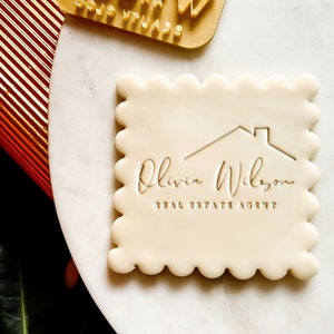 May include: A white cookie with a scalloped edge, featuring a gold embossed logo with a house icon and the text "Olivia Wilson REAL ESTATE AGENT".