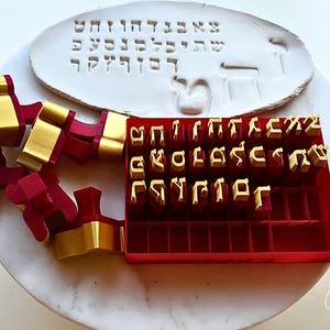 May include: A set of red and gold Hebrew alphabet stamps with a matching red storage tray. The stamps feature gold-coloured letters on red bases. An oval white clay plaque displays Hebrew characters pressed into the surface by the stamps.