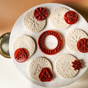 Op de afbeelding: Set van acht witte en rode koekjesvormen met Viking-symbolen, waaronder een kompas, een raaf en een knoop. De vormen zijn gerangschikt op een wit marmeren oppervlak.